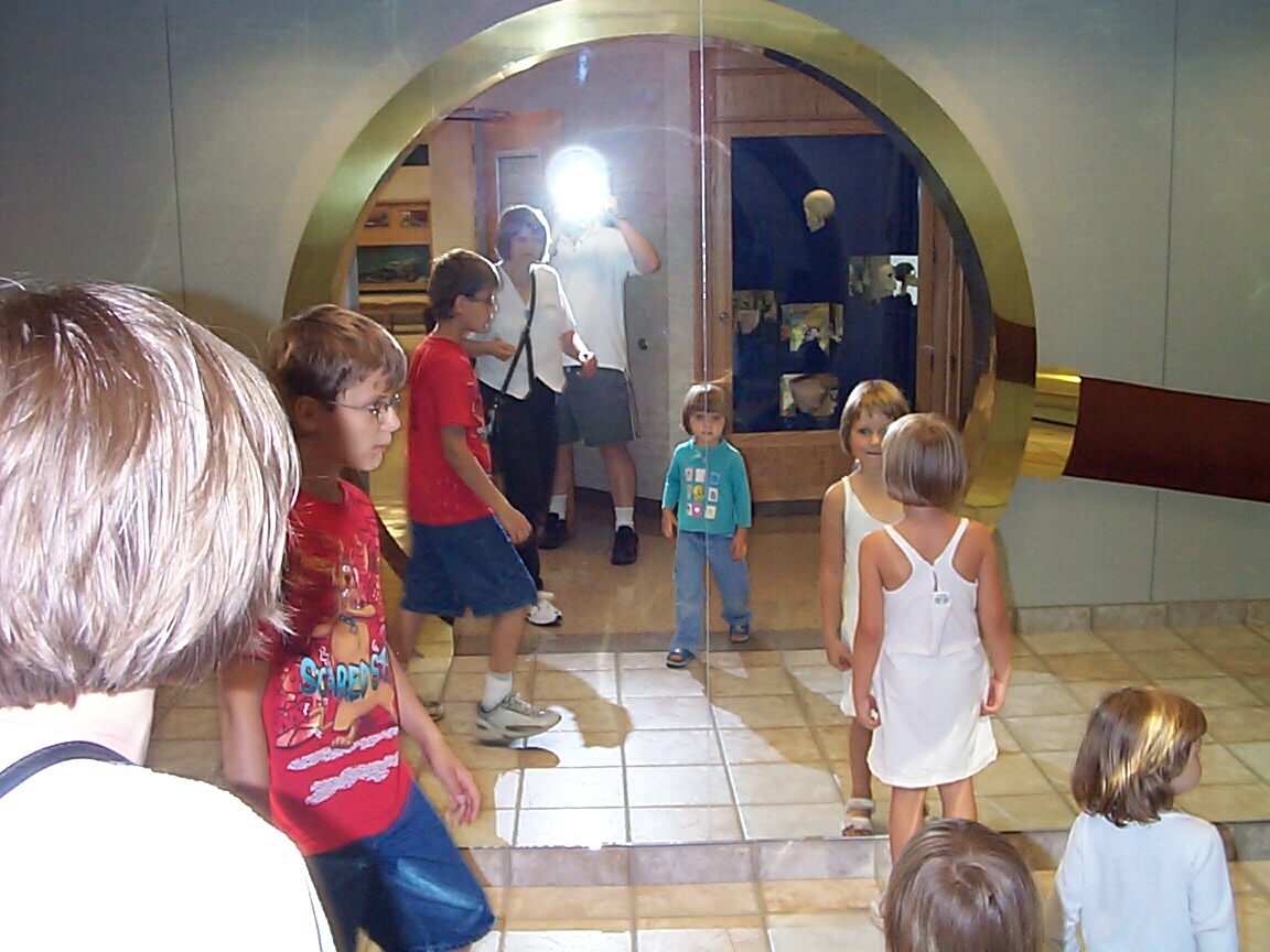 A circle of children gathered before a large round brass-framed mirror in a museum; a camera flash reflects at its center. Photographed in 2002.